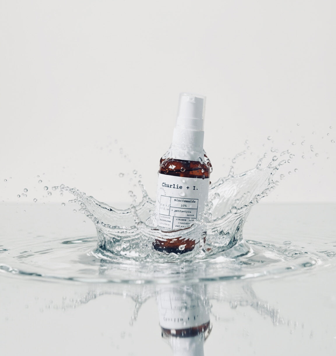 A close-up studio shot of a small, amber Charlie + I serum bottle with a white pump dispenser. The bottle, labeled "niacinamide 10%", is centered in a large splash of clear water, creating a dynamic, fresh effect on a light background.